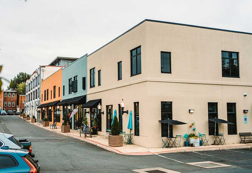 A street corner with modern beige and pastel-colored buildings housing shops and cafes. Outdoor tables with umbrellas are set up along the sidewalk. Parked cars line the street, and a mix of old and new architectural styles is visible in the background.