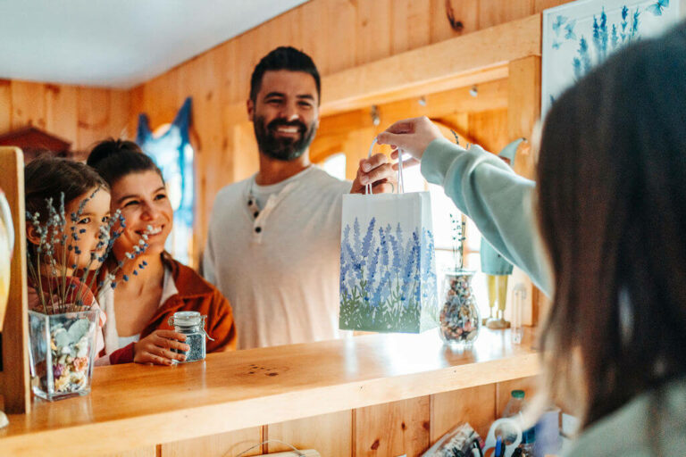 A family of three, a man, woman, and child, smile at a wooden counter inside a cozy room. Another person hands them a white bag with blue flowers. Various decorative items like jars and vases filled with flowers are arranged on the counter and shelves.