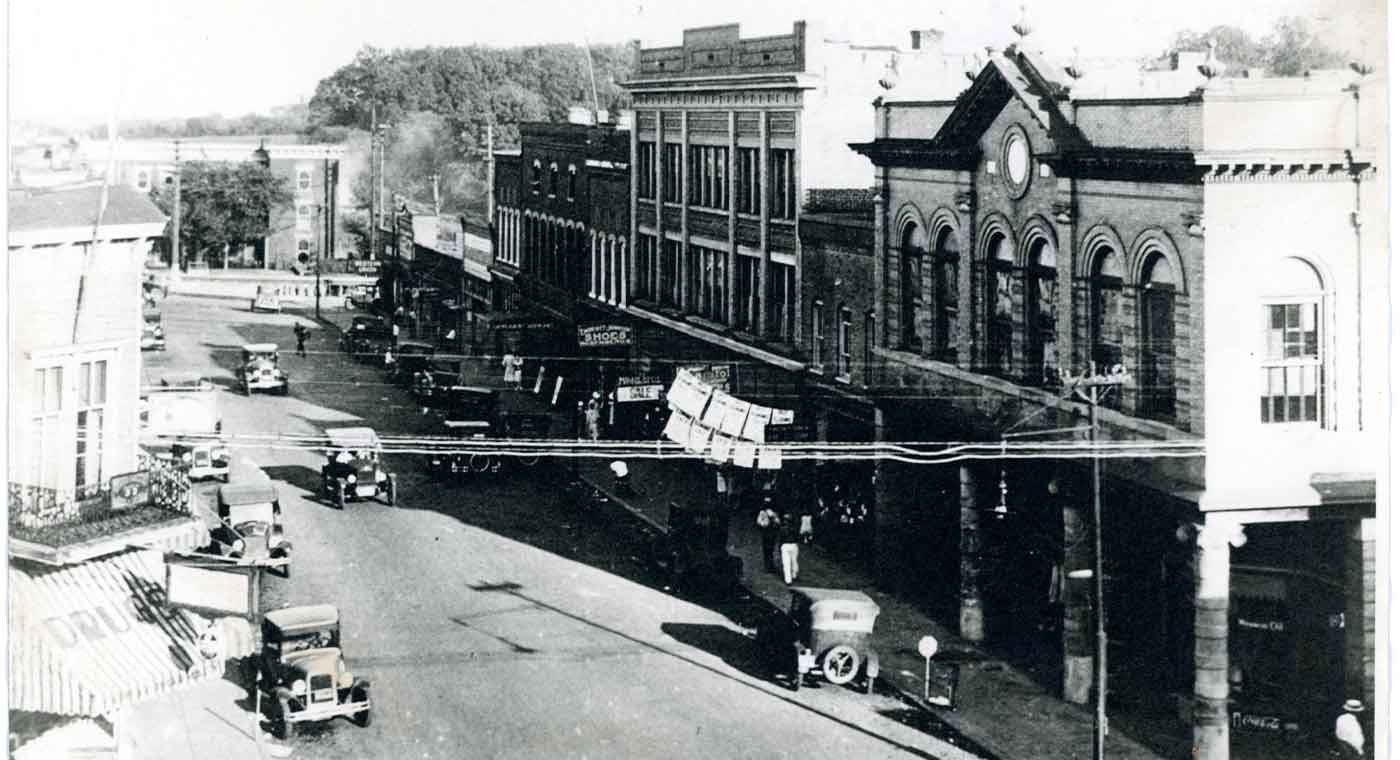 Black and white photo of a 1920s town street, featuring old automobiles, storefronts, and pedestrians. The architecture includes prominent brick buildings with ornate details. Overhead utility lines and a shaded sidewalk add to the vintage atmosphere.