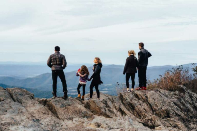 Five people, including three adults and two children, stand on a rocky outcrop overlooking a scenic mountain range. They face away from the camera, taking in the expansive view under a cloudy sky. Some wear jackets and hiking shoes, indicating a outdoor adventure.