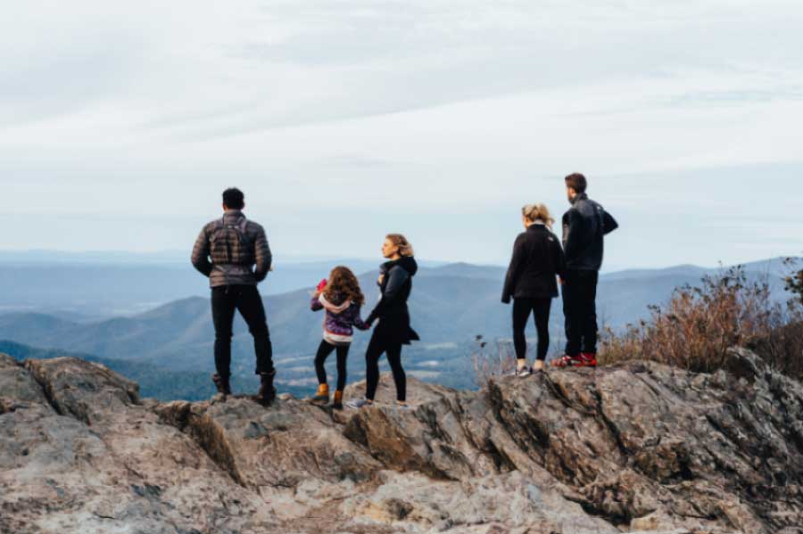 Five people, including three adults and two children, stand on a rocky outcrop overlooking a scenic mountain range. They face away from the camera, taking in the expansive view under a cloudy sky. Some wear jackets and hiking shoes, indicating a outdoor adventure.