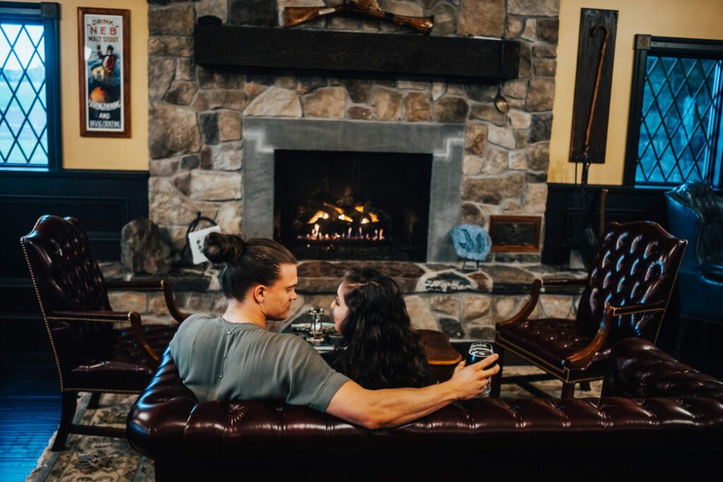 A couple with drinks sits on a brown leather couch, facing a stone fireplace with a warm fire. The rustic room has dark wooden panels, framed windows, and a poster on the wall. They appear cozy and comfortable in the homey setting.