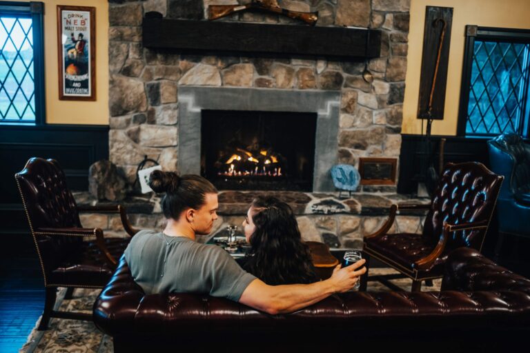 A couple with drinks sits on a brown leather couch, facing a stone fireplace with a warm fire. The rustic room has dark wooden panels, framed windows, and a poster on the wall. They appear cozy and comfortable in the homey setting.