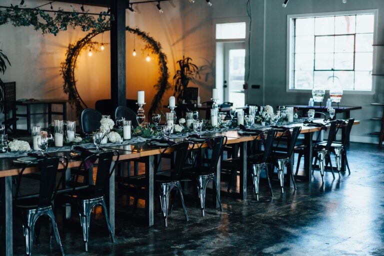 A rustic, elegantly decorated dining area with two long wooden tables set with candles, flowers, glassware, and place settings. Metal chairs provide seating, and an arch adorned with greenery and hanging lights creates a cozy ambiance in the background.
