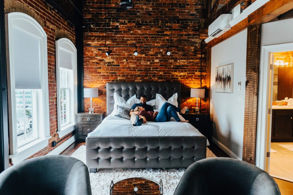 A stylish, modern bedroom with exposed brick walls features a couple lounging on a large, tufted bed with gray bedding, perfect for a weekend getaway. Two nightstands with lamps flank the bed. Large windows with blinds and a door to a brightly lit bathroom are visible in the background.