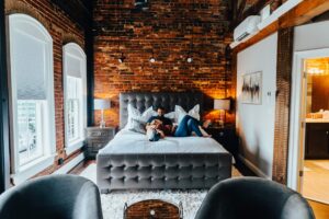 A stylish, modern bedroom with exposed brick walls features a couple lounging on a large, tufted bed with gray bedding, perfect for a weekend getaway. Two nightstands with lamps flank the bed. Large windows with blinds and a door to a brightly lit bathroom are visible in the background.