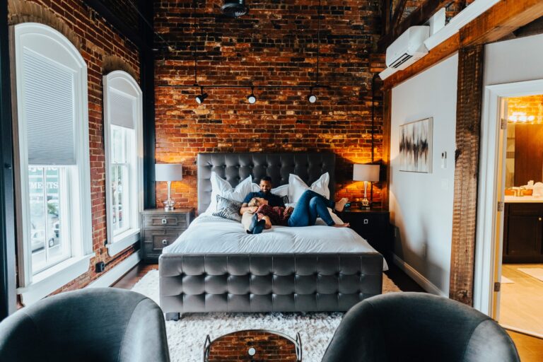 A stylish, modern bedroom with exposed brick walls features a couple lounging on a large, tufted bed with gray bedding, perfect for a weekend getaway. Two nightstands with lamps flank the bed. Large windows with blinds and a door to a brightly lit bathroom are visible in the background.