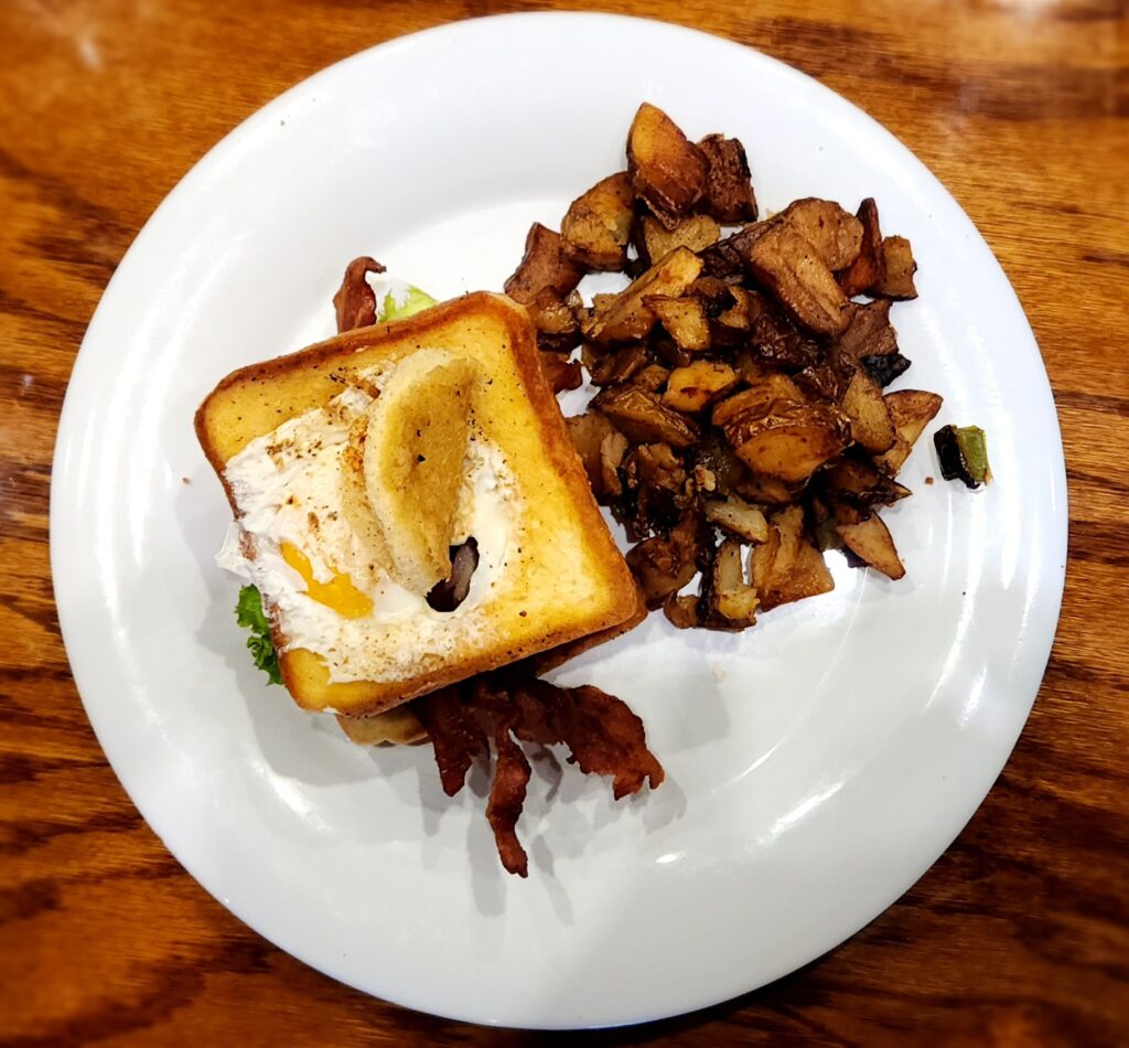 A plate sits on a wooden table, containing a brunch sandwich with egg, lettuce, and bacon in toasted bread. Next to the sandwich are seasoned, crispy roasted potatoes. The meal is presented on a white plate.