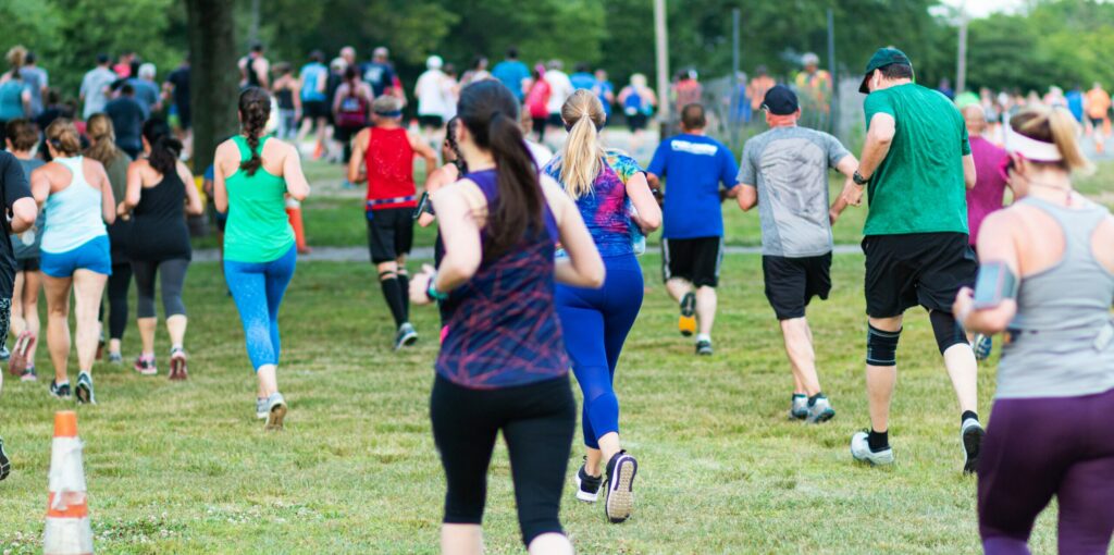 A group of people running outdoors on a grassy field, participating in a race or exercise event. They wear a variety of athletic clothing, including tank tops, shirts, and leggings. Surrounded by trees, the runners reconnect with nature as the mood remains energetic and lively.