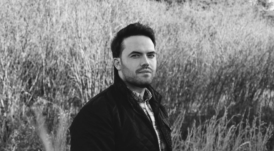 A black-and-white image of a man with short dark hair and a trimmed beard standing in a grassy field. He is wearing a plaid shirt and a quilted jacket, looking off to the side with a neutral expression. The background is dense with tall, dry vegetation.