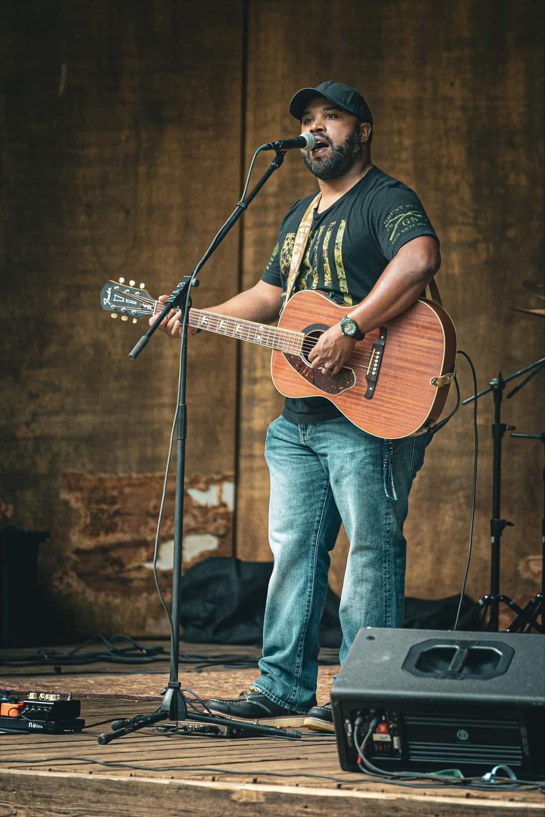 A man wearing a black cap and a black t-shirt plays an acoustic guitar and sings into a microphone. He is standing on a wooden stage with audio equipment around him. The background is a plain brown backdrop.