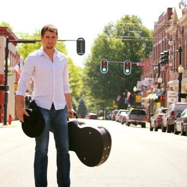 A man in a white shirt and blue jeans stands in the middle of a sunny street holding a black hat in one hand and a guitar case in the other. A row of buildings and traffic lights are visible in the background.