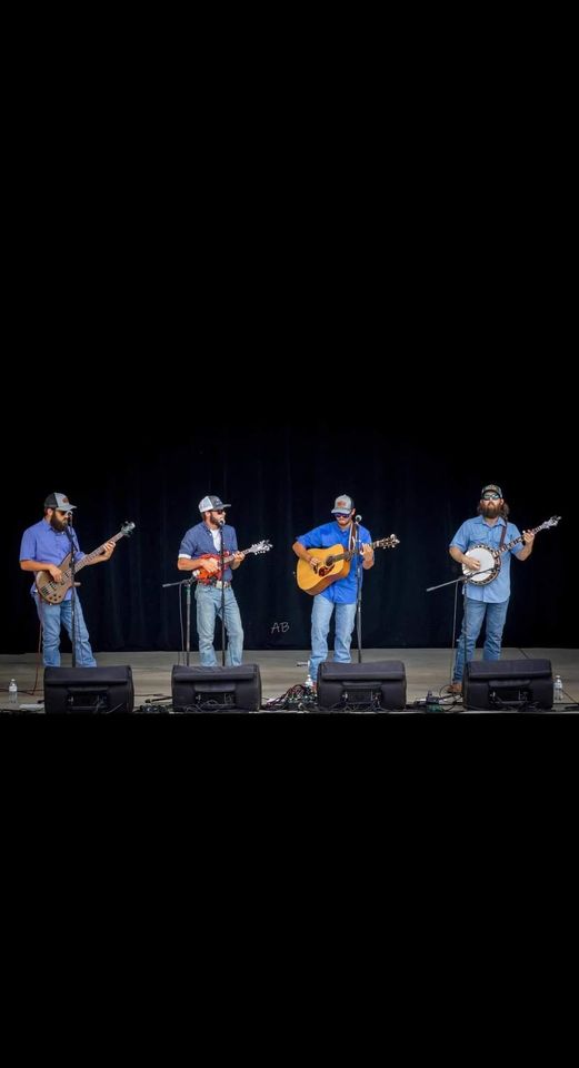 Four musicians stand on stage, each playing an instrument: a mandolin, a mandolin, a guitar, and a banjo. They wear casual attire with baseball caps and appear to be performing under stage lights with microphones in front of them.
