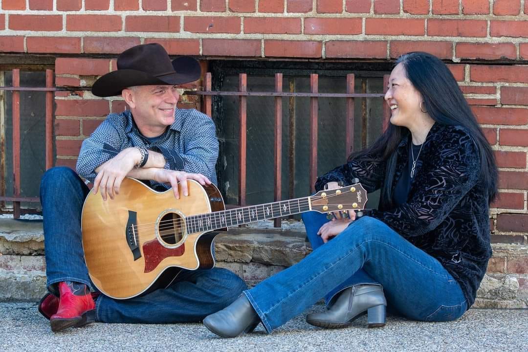 A man and woman sit on the ground against a brick wall, smiling at each other. The man, wearing a cowboy hat, holds an acoustic guitar. The woman, with long dark hair, is wearing a dark jacket and boots. It feels like Fiesta Friday! as they casually enjoy this relaxed moment together.