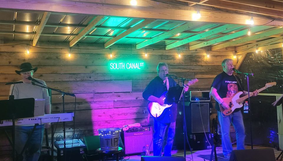 Three musicians are performing on an outdoor stage with wooden walls and string lights. The band consists of a keyboard player, a guitarist with a microphone, and a bassist. Behind them, a neon sign reads "South Canal St." The atmosphere is lively and colorful.