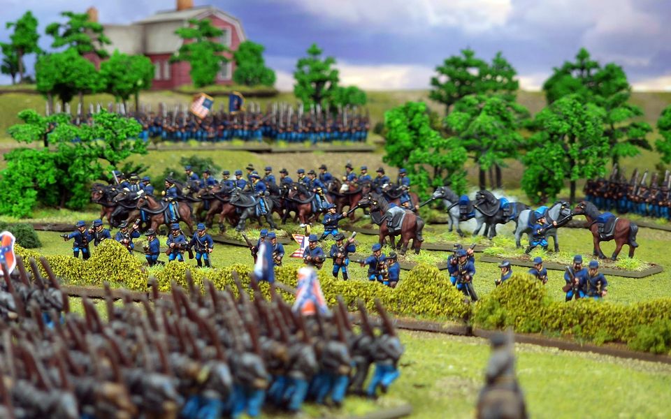 A detailed miniature diorama depicting a Civil War battlefield. Soldiers, dressed in blue uniforms, stand in formations and ride horses amidst lush green trees. A red farmhouse sits on a hill in the background. The scene is vibrant and meticulously crafted.