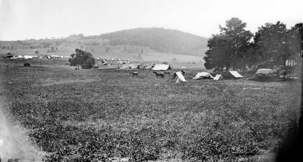 A black and white photo shows a vast field with several tents and horses scattered throughout. Trees are clustered on the right side, and a hill with additional tents lies in the background. The atmosphere resembles a historical military encampment.