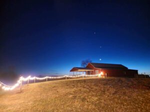 A red barn sits on a grassy hill under a deep blue evening sky. The barn is brightly lit, and a string of white lights runs along a fence leading up to it. The sky features a crescent moon and two bright, aligned stars.